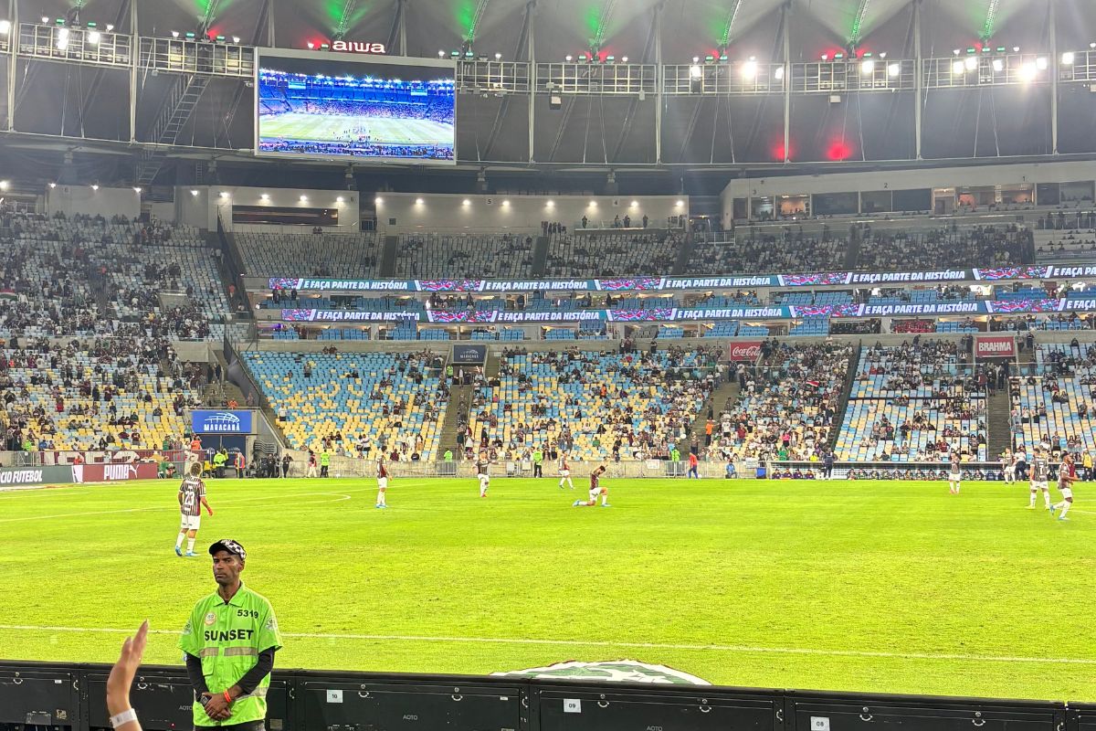 Jogadores do Fluminense em campo no Maracanã vistos da beira do gramado, com arquibancadas e telão ao fundo