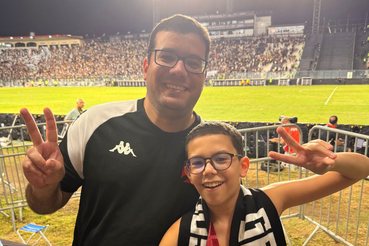 Pai e filho torcedores do Vasco sorrindo e fazendo sinal de vitória com o gramado de São Januário ao fundo